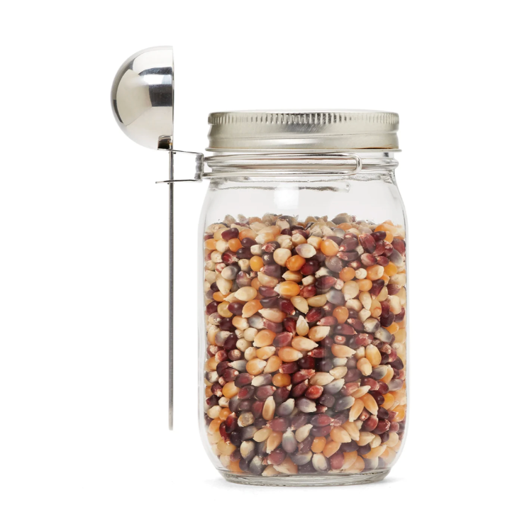 Un pot en verre contenant des grains de popcorn avec une cuillère en acier inoxydable attaché sur le pot sur un fond blanc