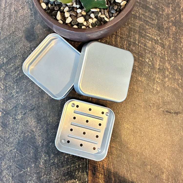 Les 3 étages d'un boitier de métal carré en aluminium pour shampoing solide permettant un drainage du shampoing en barre. Arrière plan d'une planche en bois et d'une plante succulente.