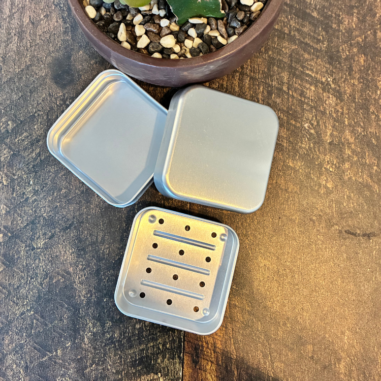 Les 3 étages d'un boitier de métal carré en aluminium pour shampoing solide permettant un drainage du shampoing en barre. Arrière plan d'une planche en bois et d'une plante succulente.