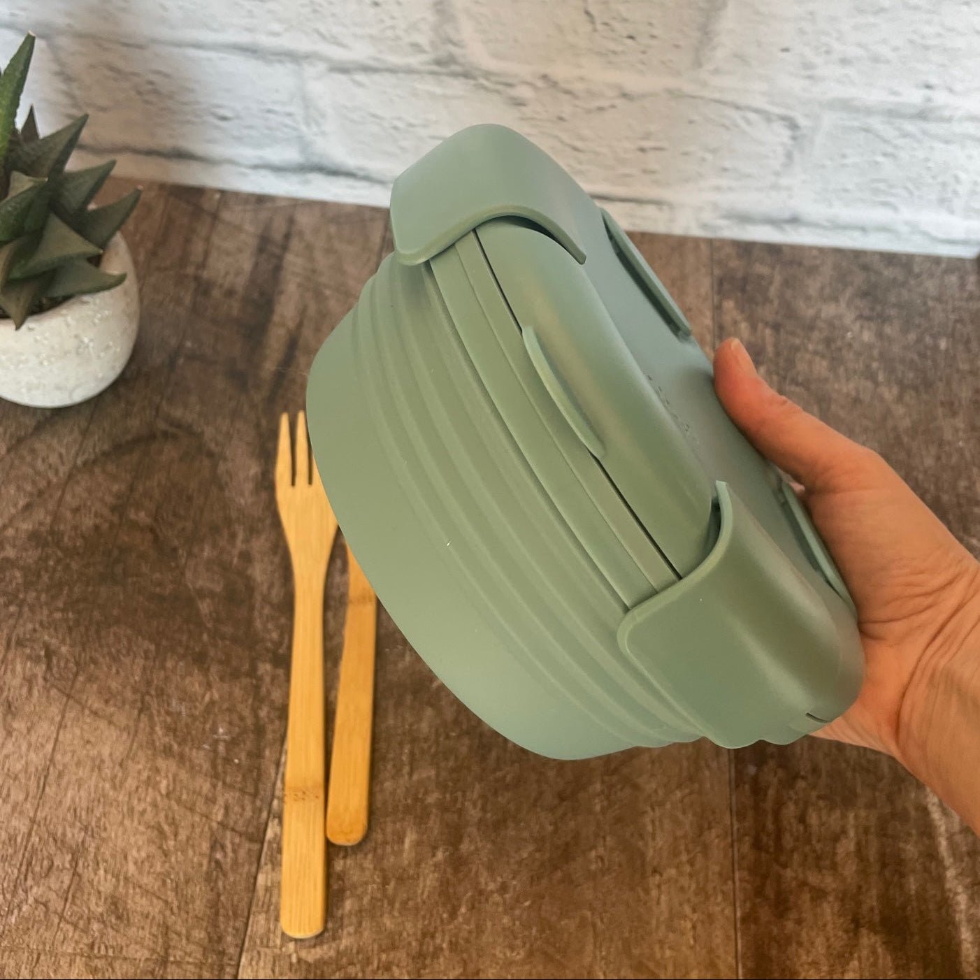 bol en silicone rétractable en plein dimension de couleur eucalyptus avec un couvercle comprenant 4 clips sur le couvercle tenu dans une main et un arrière plan de brique blanche.