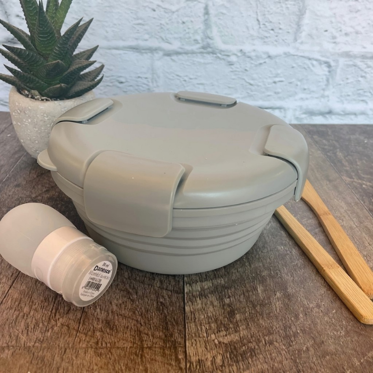 bol en silicone rétractable de couleur gris avec un couvercle comprenant 4 clips sur le couvercle déposé sur une blanche de bois avec des ustensiles en bois, une petite plante et un arrière plan de brique blanche.