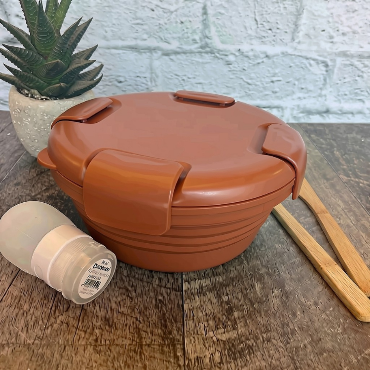 bol en silicone rétractable de couleur rouille avec un couvercle comprenant 4 clips sur le couvercle déposé sur une blanche de bois avec des ustensiles en bois, une petite plante et un arrière plan de brique blanche.