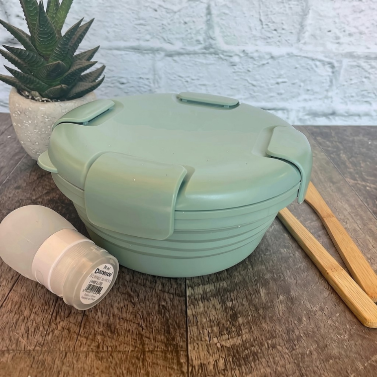 bol en silicone rétractable de couleur sauge avec un couvercle comprenant 4 clips sur le couvercle déposé sur une blanche de bois avec des ustensiles en bois, une petite plante et un arrière plan de brique blanche.