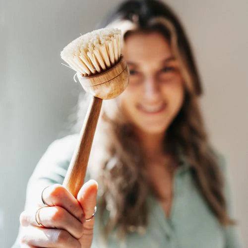 Jeune femme qui sourie hors focus flou qui tient une brosse à vaisselle en bois devant la caméra en gros plan. fond blanc gris
