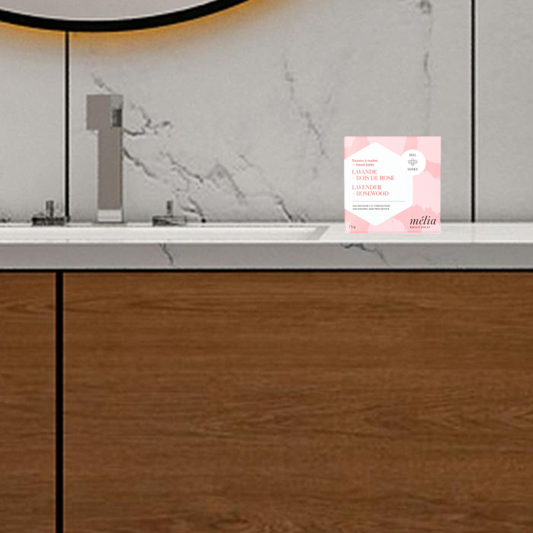 Petite boite rose sur laquelle il est écrit Mélia baume à main déposée sur un comptoir de salle de bain avec une vanité en bois.