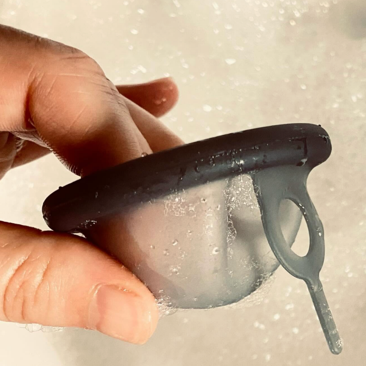 Une disque menstruelle avec languette noire tenu dans une main dans une eau savonneuse.