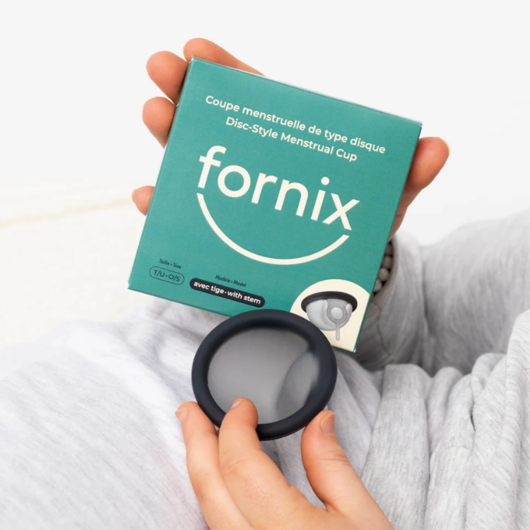 Une boite bleu avec l'écriture coupe menstruelle de type disque Dis style menstrual Cup Fornix et un disque noir en silicone tenu dans des mains de femme avec un fond blanc.