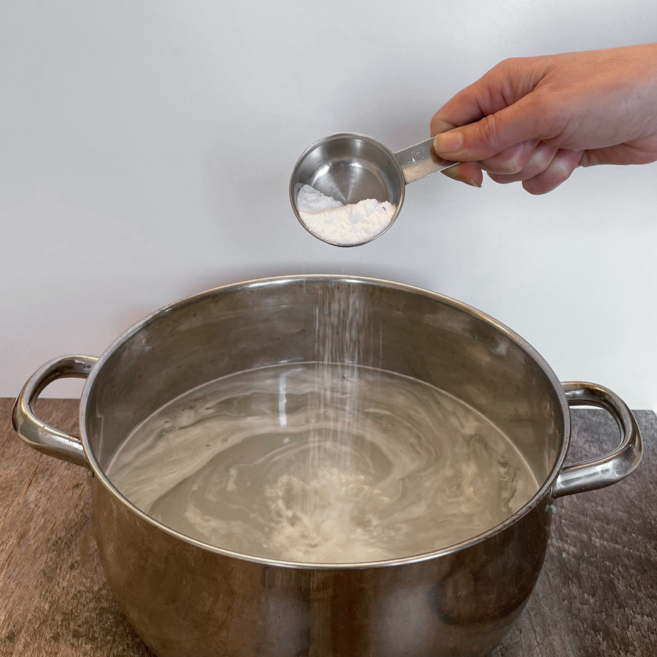 Main qui verse de la poudre blanche dans une tasse à mesurer en métal dans de l'eau contenu dans un chaudron de métal sur un comptoir de bois avec un fond blanc
