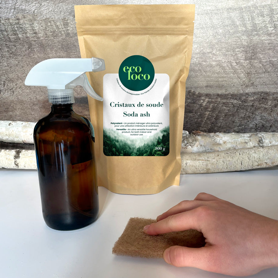 Pochette en papier kraft Eco Loco de cristaux de soude sur un fond de mur de bois pâle avec des branches de bouleaux, un comptoir blanc, une bouteille en verre brune foncée et une main qui frotte le comptoir avec une éponge brune