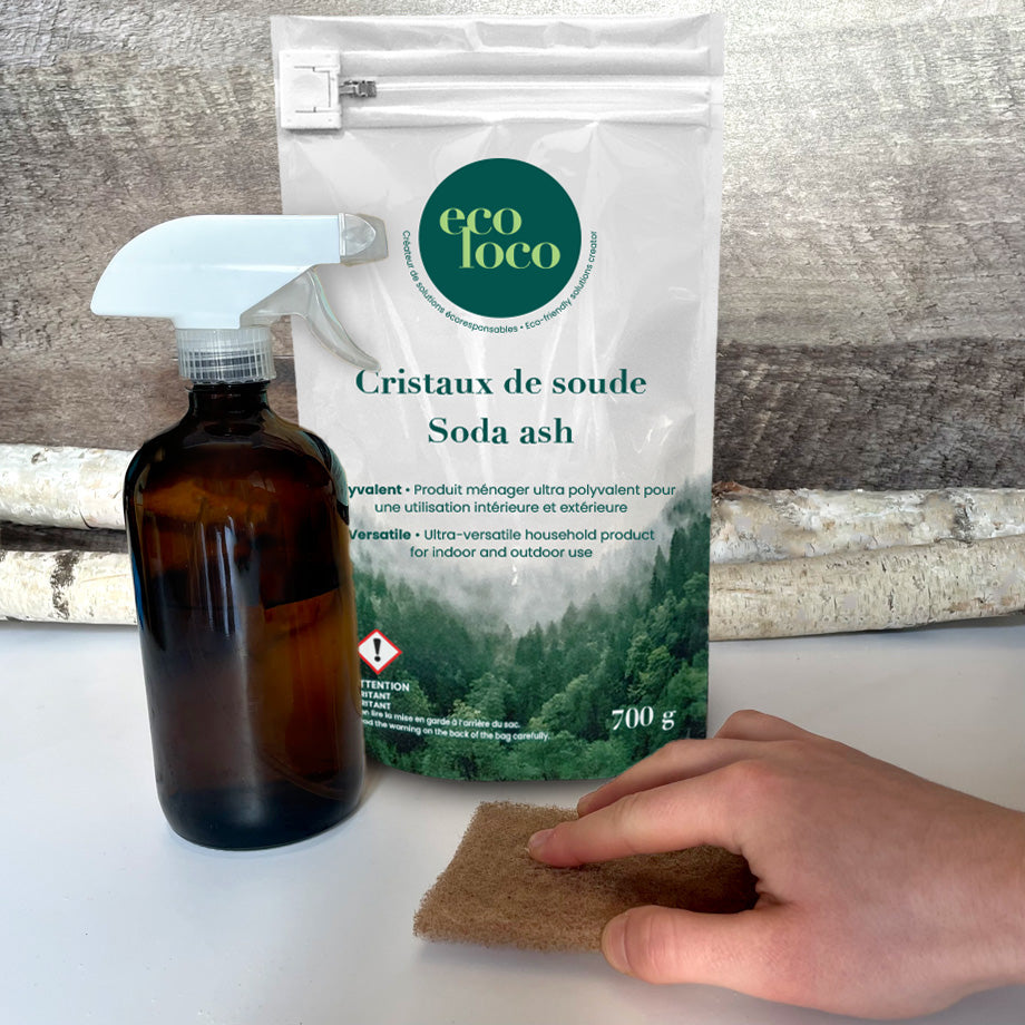 Pochette en Eco Loco de cristaux de soude sur un fond de mur de bois pâle avec des branches de bouleaux, un comptoir blanc, une bouteille en verre brune foncée et une main qui frotte le comptoir avec une éponge brune