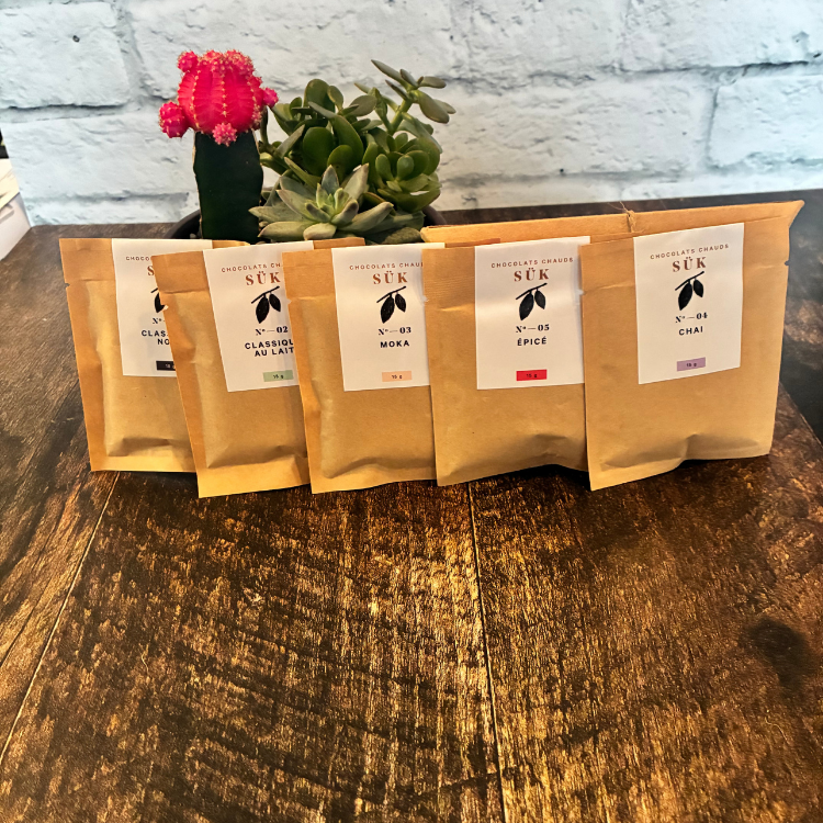 Les 5 saveurs de chocolat chauds en sachet kraft de la compagnie SÜK adossé à une plante sur une planche de bois.