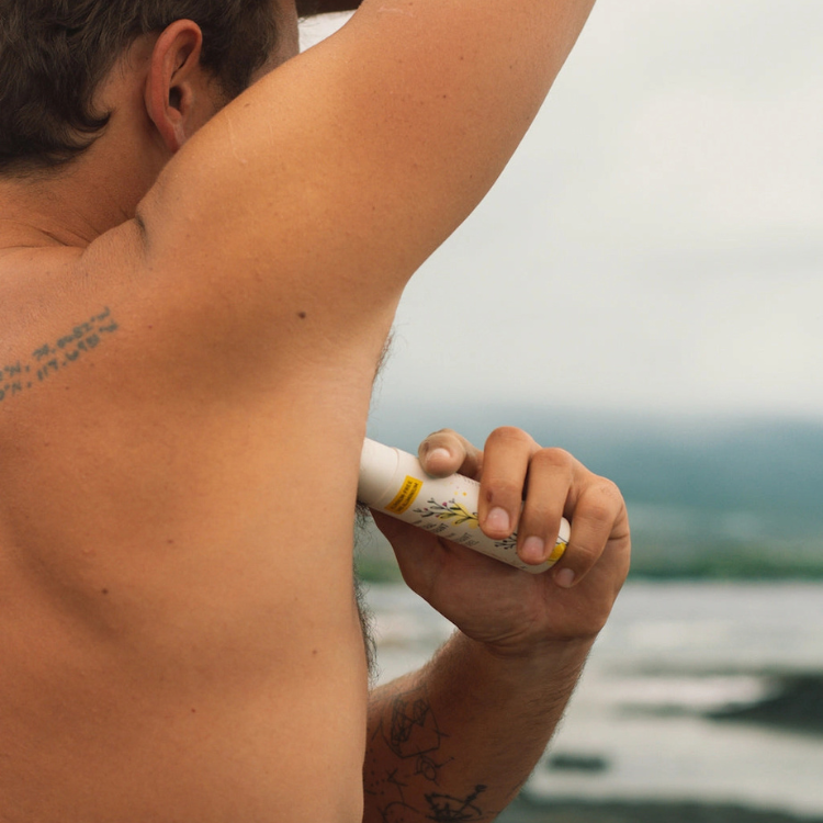 Un homme avec un tatoo qui se met du déodorant avec un fond de mer et de ciel bleu.