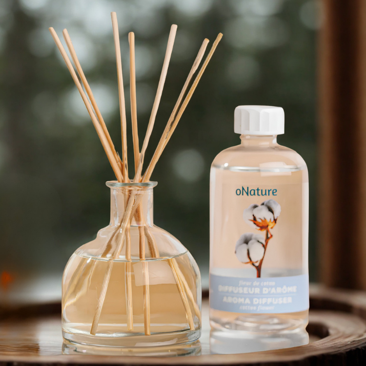 Une bouteille en verre contenant des batonnet en bois et ayant à sa droite une bouteille sur laquelle on voit une fleur de cotton et les mots: Diffuseur d'arôme, aroma diffuseur, fleur de cotton.