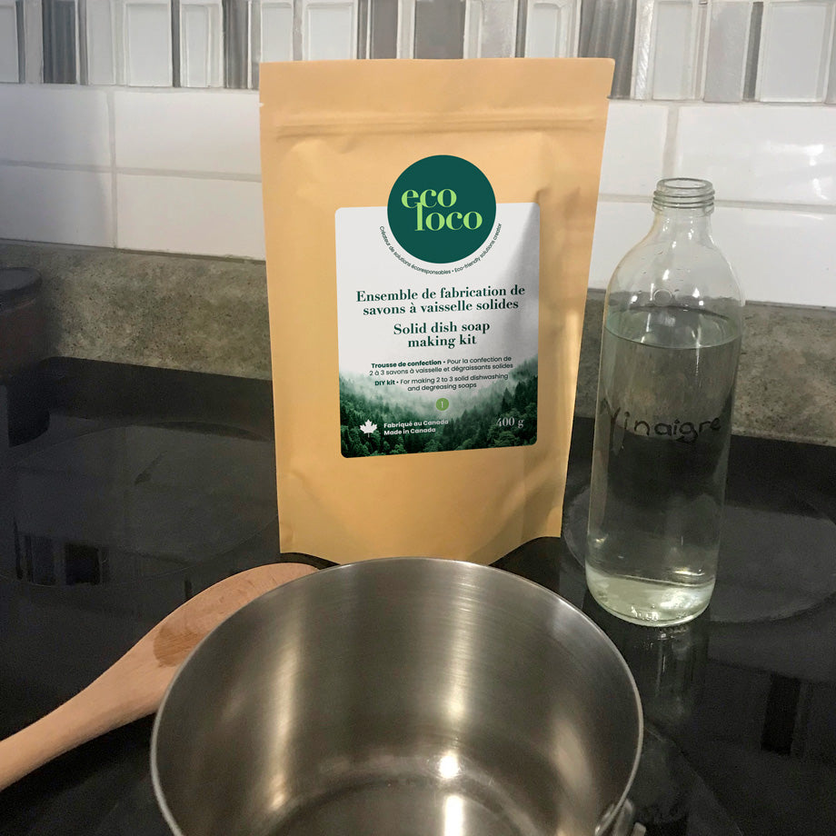 Pochette en papier kraft avec le logo d'Eco Loco contenant un ensemble de fabrication de savons à vaisselle solides sur une comptoir de cuisine avec un chaudron en métal, une bouteille en verre vitre contenant du vinaigre et une cuillère en bois