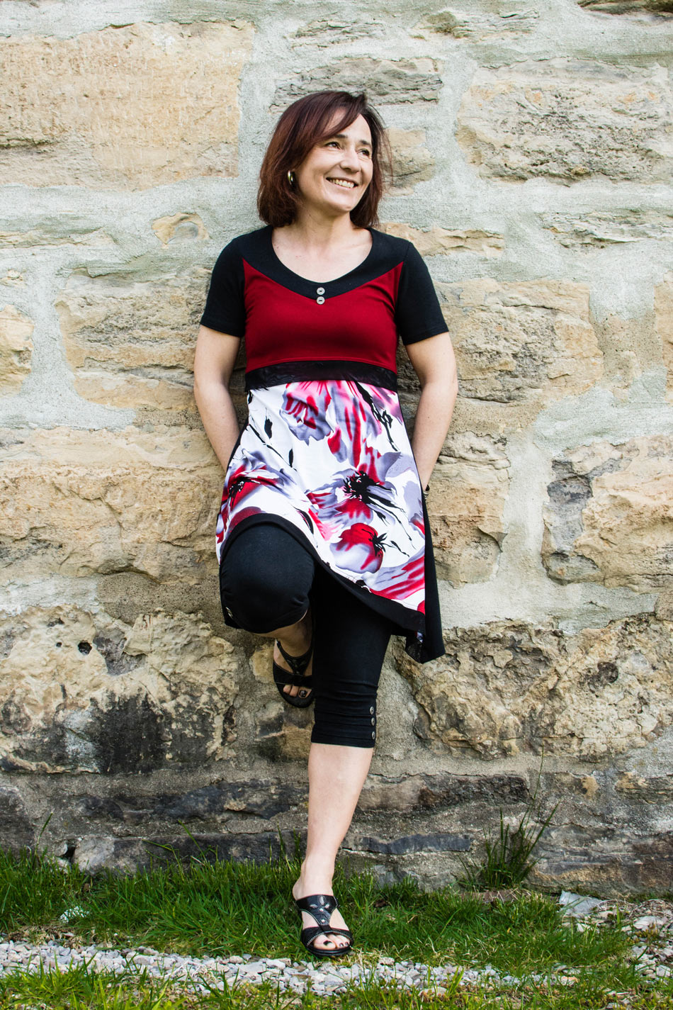 Femme souriante Geneviève St-Amour qui se tient appuyée sur un mur de vieilles pierres beige portant une robe rouge fleurie | ecoloco