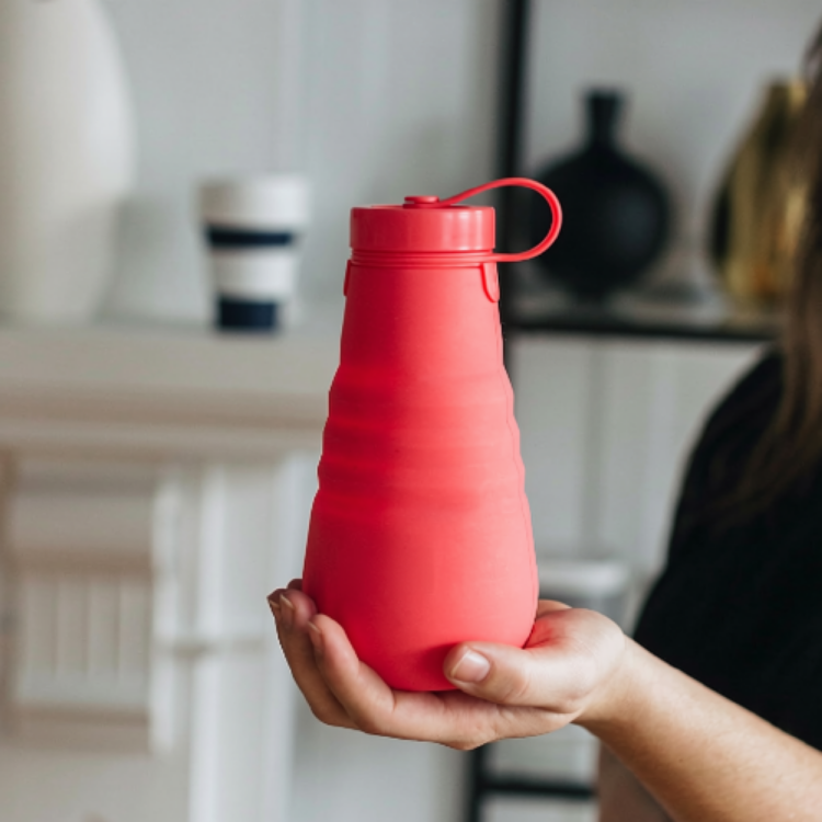 Une gourde rouge en silicone avec une ganse tenu dans une main féminine avec un fond flou majoritairement blanc.