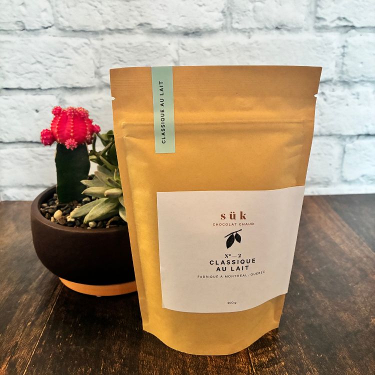 Sac de chocolat chaud classique au lait de SÜK une entreprise québécoise sur une planche de bois avec une plante en arrière plan.