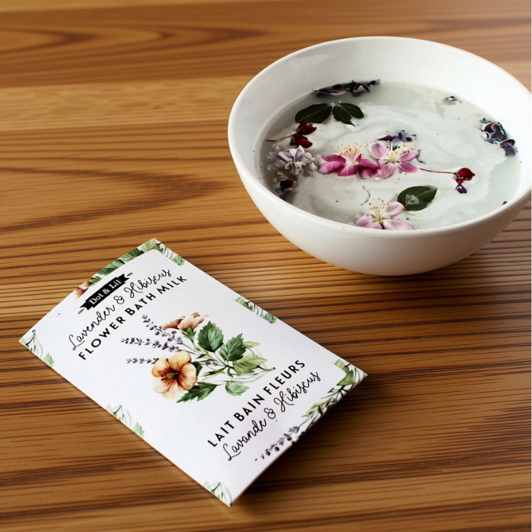 Une sachet rectangulaire blanc écrit lait de bain fleure lavande et hibiscus en francais et en anglais et le logo de Dot & Lil avec un bol d'un liquide laiteux avec de fleur sur un fond de planche en bois.