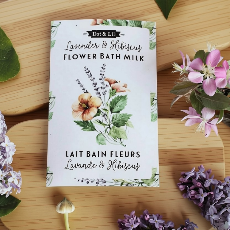 Une sachet rectangulaire blanc écrit lait de bain fleurs Lavande et hibiscus en francais et en anglais et le logo de Dot & Lil sur une planche en bois, avec une des lilas et autre type de fleurs.