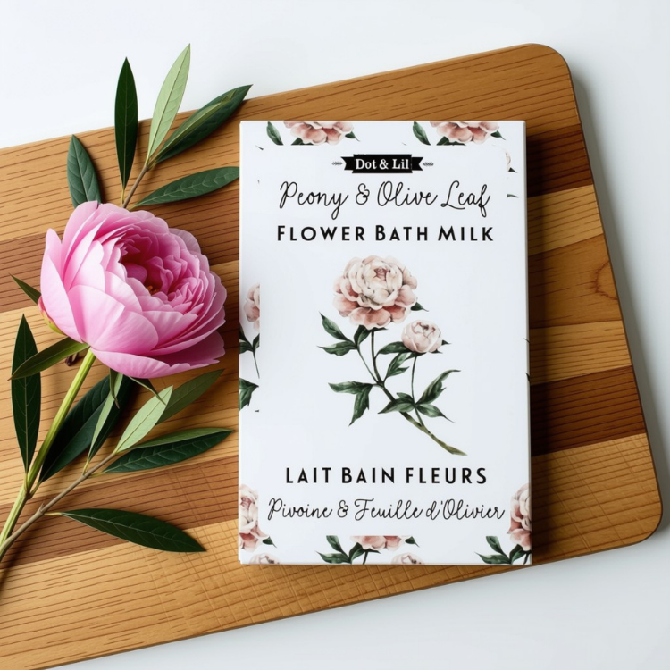 Une sachet rectangulaire blanc écrit lait de bain fleure pivoine et feuille d'olivier en francais et en anglais et le logo de Dot & Lil sur une planche en bois, avec une pivoine rose et des feuilles d'olivier sur fond blanc.