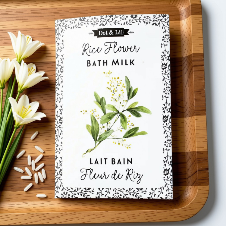 Une sachet rectangulaire blanc écrit lait de bain fleur de riz en francais et en anglais et le logo de Dot & Lil sur une planche en bois, avec des fleurs de riz et sur fond blanc.