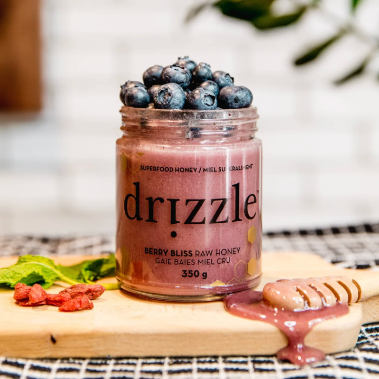 Pot de miel rose Drizzle Berry bliss de 350 grammes avec des bleuets sur le dessus du pot, des baie de gogie, une cuiller en bois et du miel coulant déposé sur une planche en bois.