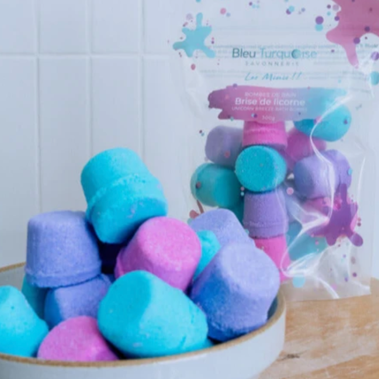 Mini bombes de bain en forme de bouchon multicolores bleu, rose et mauve dans un pot blanc avec un sac de Bleu Turquoise savonnerie en arrière plan.