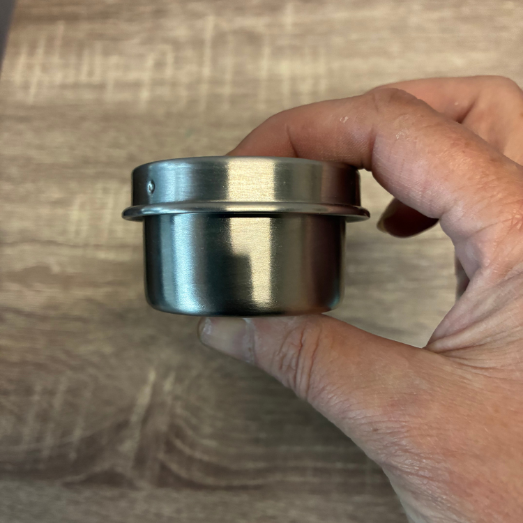 Un petit pot avec un couvercle en acier inoxydable argent enu dans une ain de femme au-dessus d'une planche en bois.