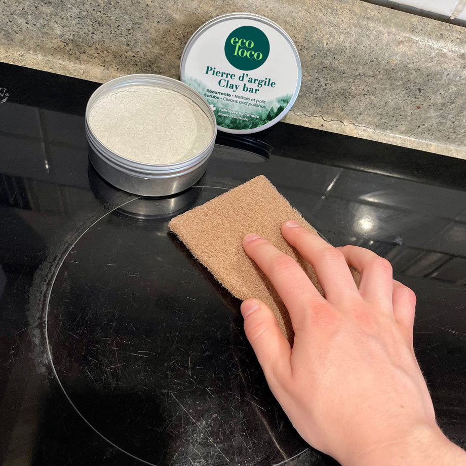 Main qui lave un dessus de four en vitrocéramique avec une pierre d'argile fabriquée par Eco loco et une éponge en fibre de noix naturelle