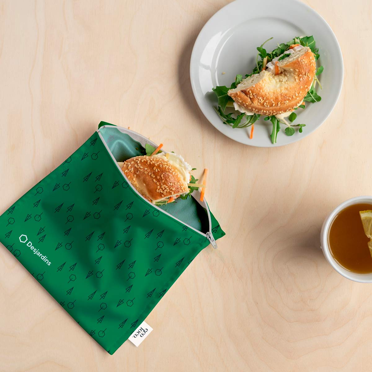 Un sac réutilisable format sandwich vert et logo Desjardins contenant un demi bagel assiette avec bagel tasse de thé avec tranche de citron sur fond texturé de bois