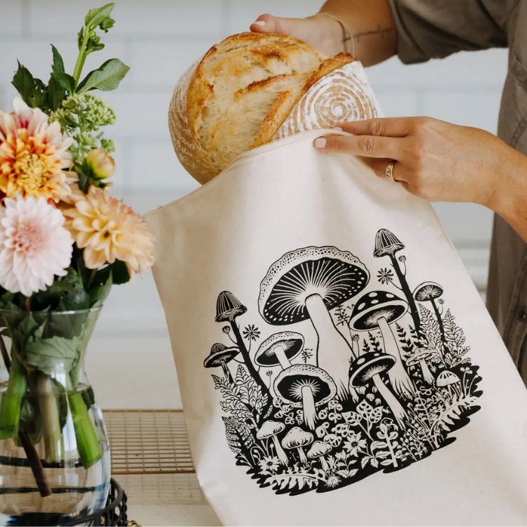 Une main qui met une miche de pain dans un sac à pain réutilisable de couleur naturelle avec des motifs de champignons. À gauche du sac à pain, il y a un bouquet de fleur dans un pot en verre.