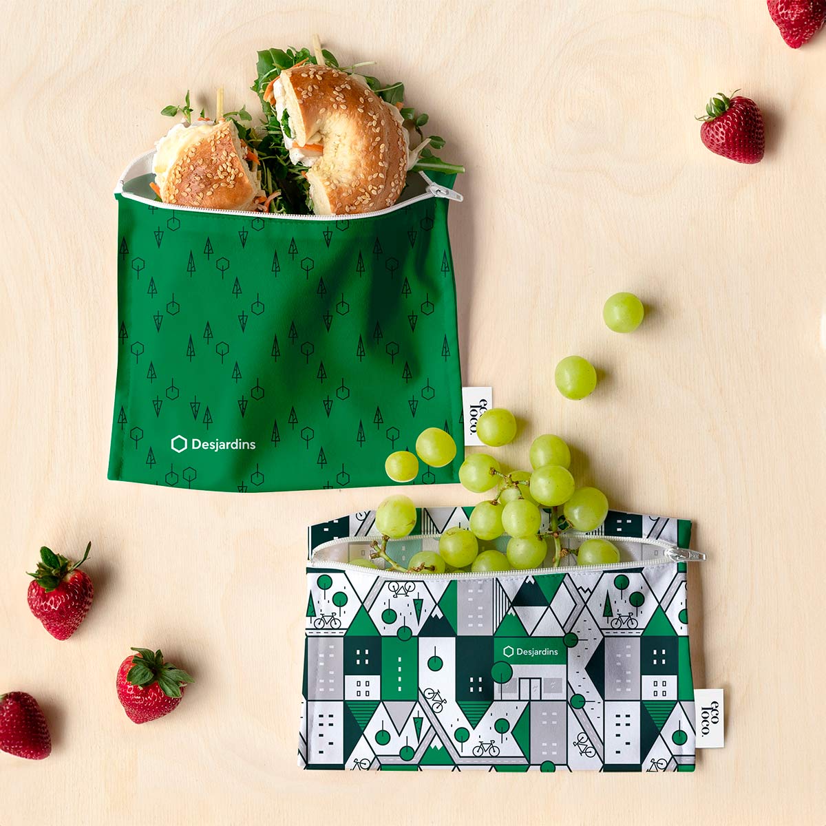 Deux sacs réutilisables aux couleurs du branding de Desjardins objets promotionnels ecolo avec à l'intérieur un bagel et des raisins des fraises sont sur le fond texture de bois