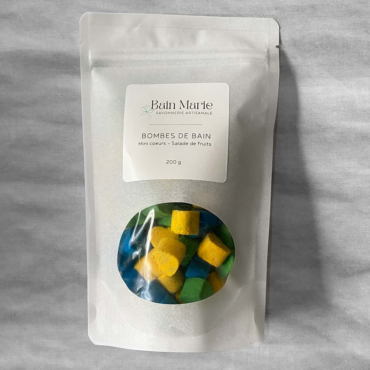 sac blanc avec une fenêtre qui laisse paraitre des petites coeursjaunes, verts et bleus sur un fond blanc effet de marbre gris. Étiquette blanche avec les inscriptions Bain Marie Savonnerie artisanale Bombes de bain mini coeurs sortilège 200g