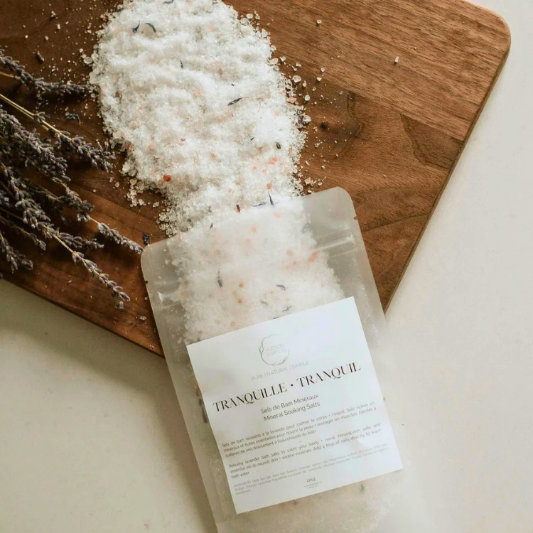 Un sac de sel de bain avec l'inscription Transquille - refresh ouvert et se déversant sur une planche de bois. Une branche de lavande se trouve sur la gauche et le fond est blanc.