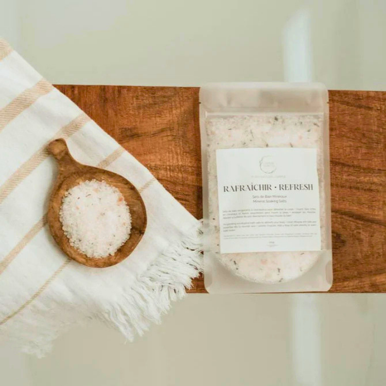 Un sac de sel de bain avec l'inscription rafraichir - refresh sur une planche de bois. À sa gauche un vois un petit récipient de sel de bain déposé sur une serviette.