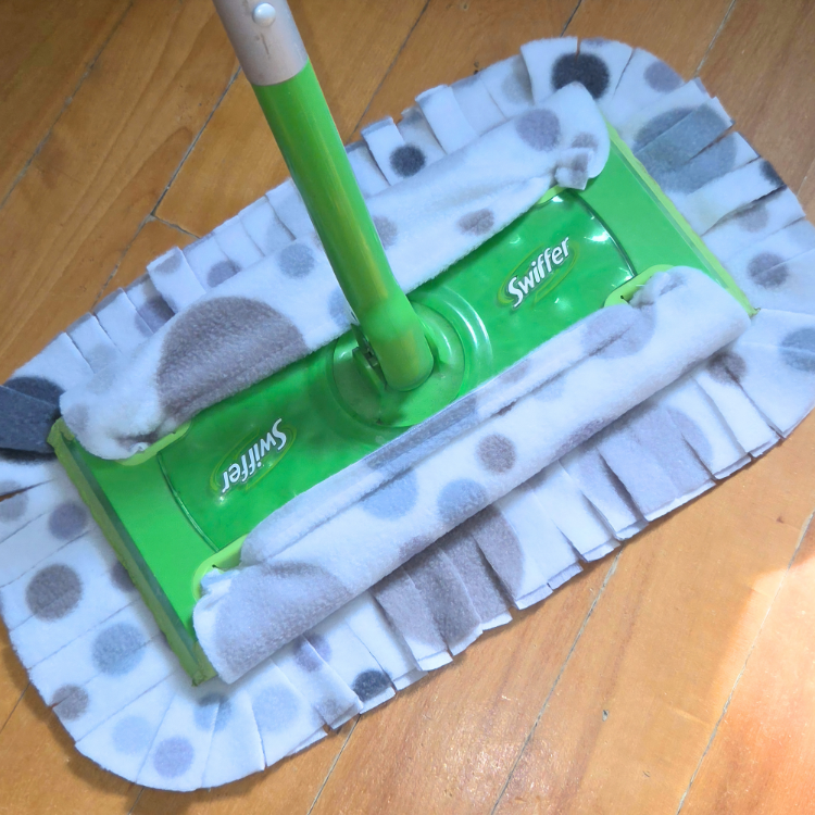 Un tissu gris pâle avec de rond gris et beige et coupé en lanière en bordure mis sur un manche à balai swiffer avec un plancher en bois en fond.