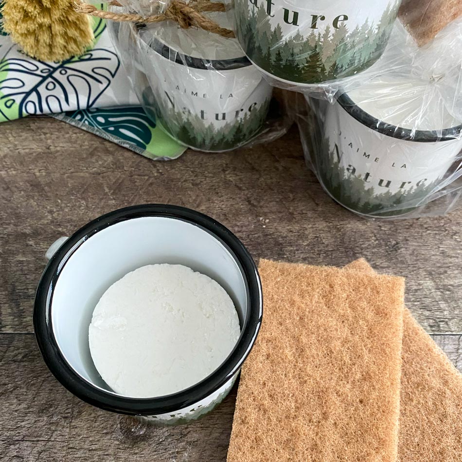 Tasses en acier émaillée sur un comptoir en bois foncé entourée de tampon à récurer en noix, brosse à vaisselle en bois et un linge à vaisselle en microfibre au motif de feuilles tropicales vertes