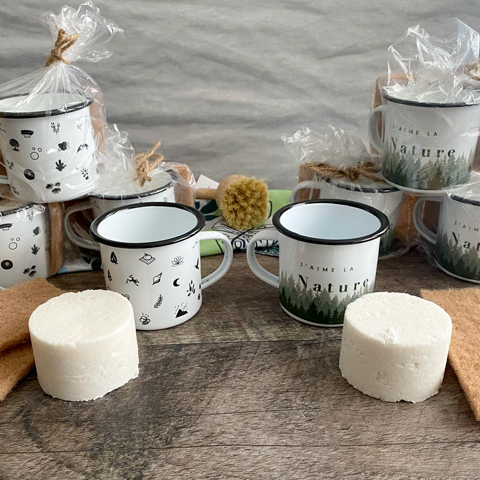 Tasses en acier émaillée sur un comptoir en bois foncé entourée de tampon à récurer en noix, brosse à vaisselle en bois et un linge à vaisselle en microfibre au motif de feuilles tropicales vertes