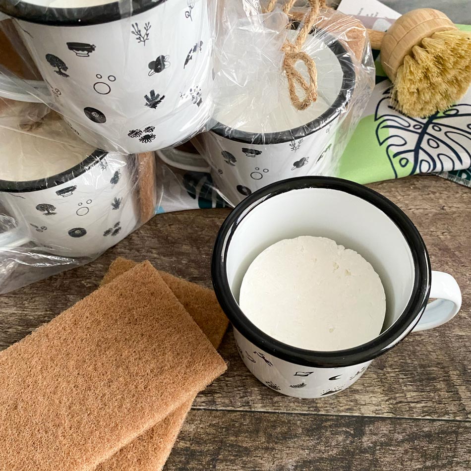 Tasses en acier émaillée sur un comptoir en bois foncé entourée de tampon à récurer en noix, brosse à vaisselle en bois et un linge à vaisselle en microfibre au motif de feuilles tropicales vertes