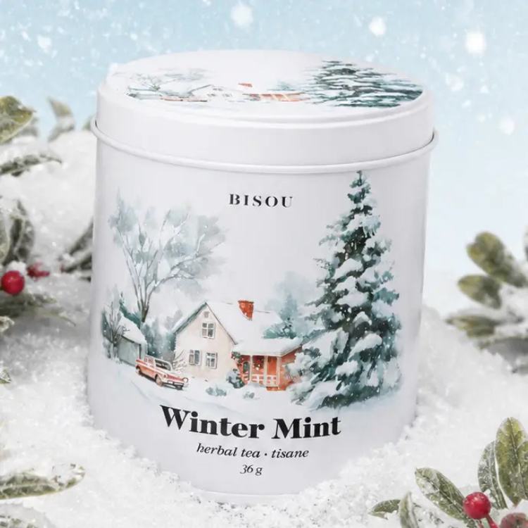 Une boite ronde et blanche avec un dessin d'un salon chaleureux comprenant une maison, une voiture et des arbres couverts de neige et des inscriptions Bisou winer mint entouré de neige et de plantes.