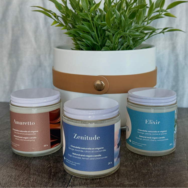 Trio de chandelle avec des étiquettes de différentes couleurs sur lesquelles on peut lire: Amaretto, Zenitude et Elixir en avant d'un pot blanc contenant une plante verte.