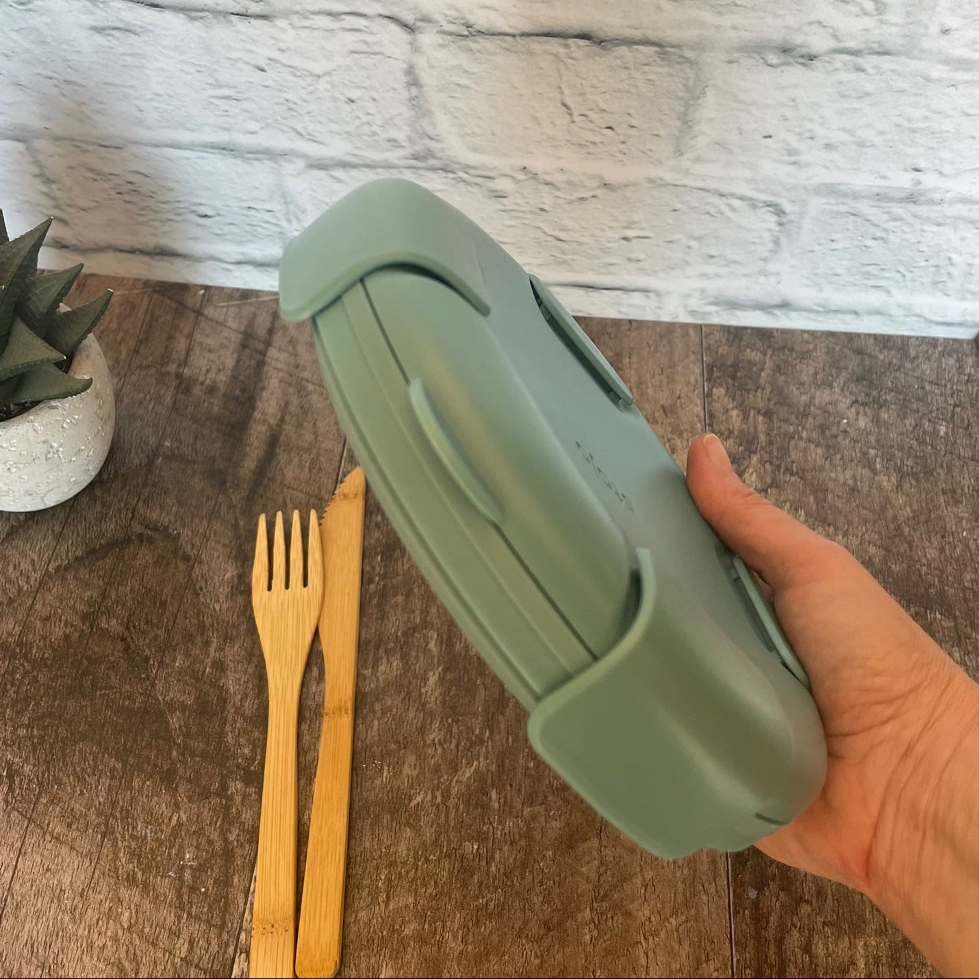 bol en silicone rétractable de couleur eucalyptus avec un couvercle tenu dans une main et un arrière plan de brique blanche.