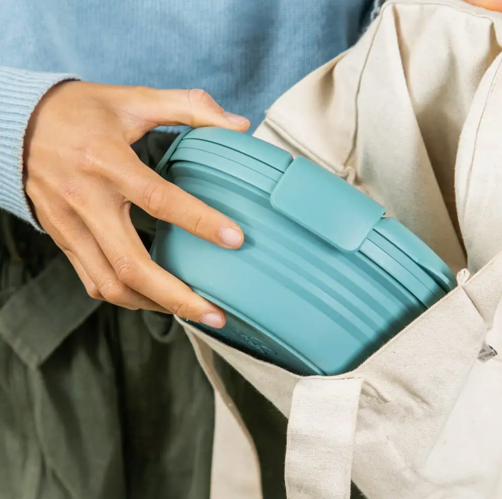 bol en silicone rétractable de couleur eucalyptus avec un couvercle mis dans un sac en coton de couleur naturelle par une main féminine