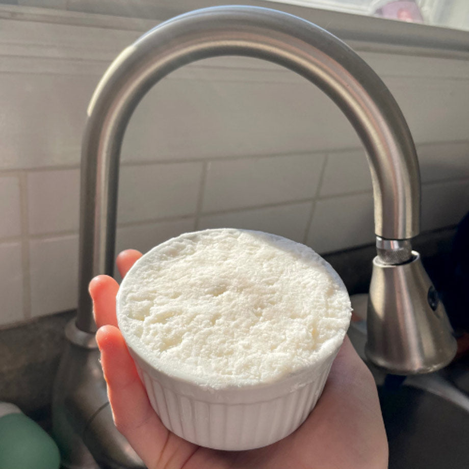 Une main qui tient un cake vaisselle fabriqué par Eco Loco avec un robinet en fond dans uen cuisine