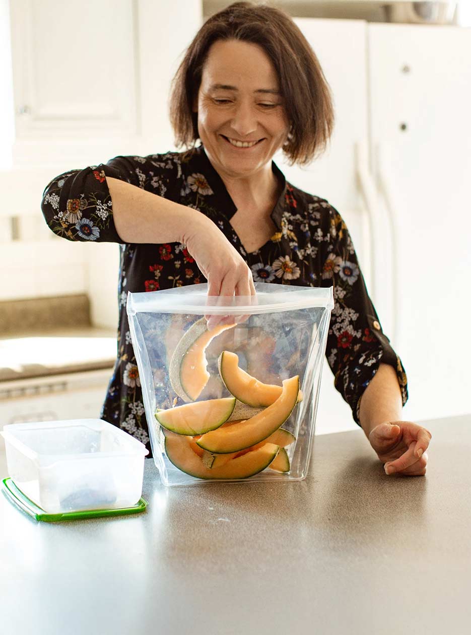 Femme aux cheveux bruns qui sourit en mettant une tranche de cantaloup dans un grand sac en silicone écoresponsable dans une cuisine blanche et lumineuse