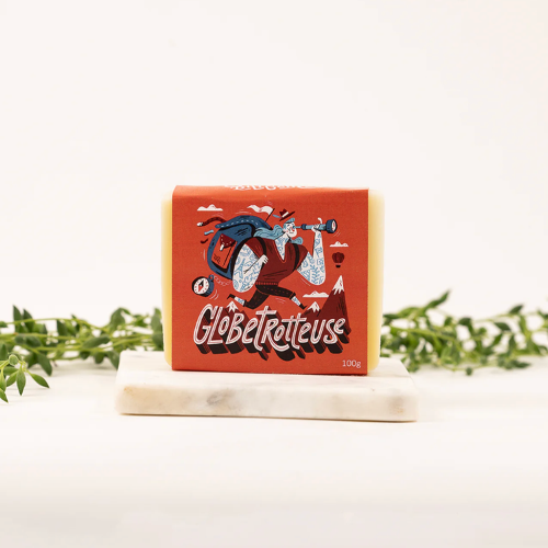 Savon la Globetrotteuse pour le corps, les cheveux et la lessive de la savonnerie des Diligences sur un fond minimaliste composé d'un porte-savon blanc et de feuille.