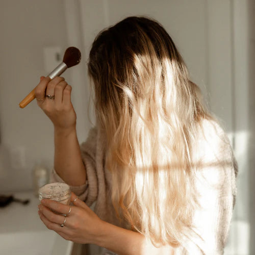 jeune femme qui tient un pinceau et qui se met du shampoing sec dans les cheveux bordé de lumière naturelle