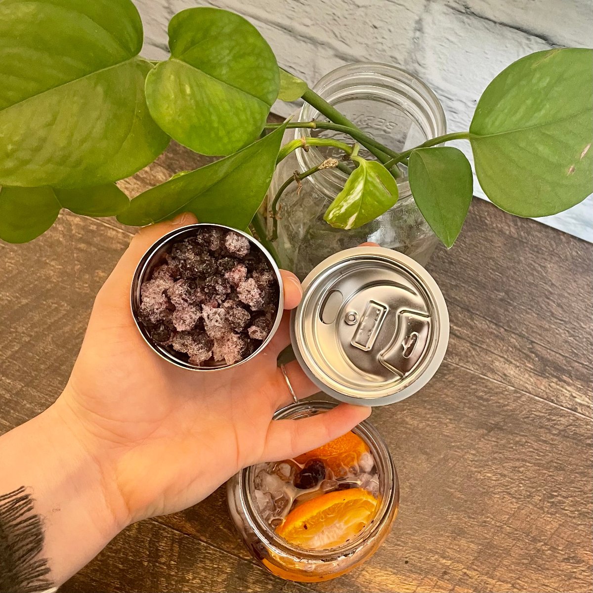 Infuseur à thé ou autre avec une main qui le tient au dessus d'un verre avec de la glace et une orage sur une table de bois avec une plante et un mur de briques blanches | ecoloco