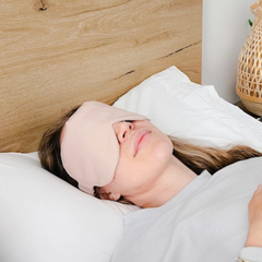 Une femme aux cheveux longs couchée sur un lit avec des draps blancs et une tête de lit en bois portant un masque pour les yeux obscurcissant aussi appelé masque de loup de couleur rose très pâle. 