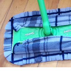 Adaptateur en tissu pour balai swiffer carroté gris sur un balai swiffer déposé sur un plancher de bois. 